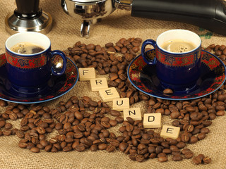 Coffee beans, two ornate cups, tamper and group handle with the letters Freunde on a hessian background