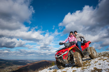 Smiling couple in winter clothes on a red quad bike on a mountain slope under the blue cloudy sky on background of mountains and the town in the valley