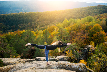 Attractive fit female is practicing yoga and doing asana Virabhadrasana 3 on the top of the mountain in the evening. Beautiful sunset, autumn forests, rocks and hills on the background