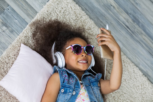 cute African girl at home, listening to music in big white headphones, Lies on the floor