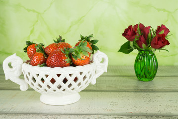Frische Erdbeeren in eine weißen Porzellanschale und rote Rosen in einer kleinen Vase