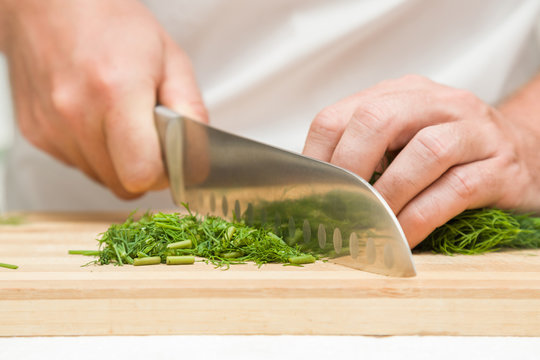 Chef's hands with knife cutting and crushing a dill on the wooden board. Preparation for cooking. Healthy eating and lifestyle.
