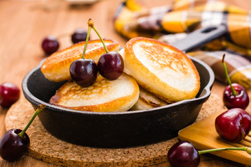 Close-up of fried pancakes in a iron pan and ripe cherries on a wooden table
