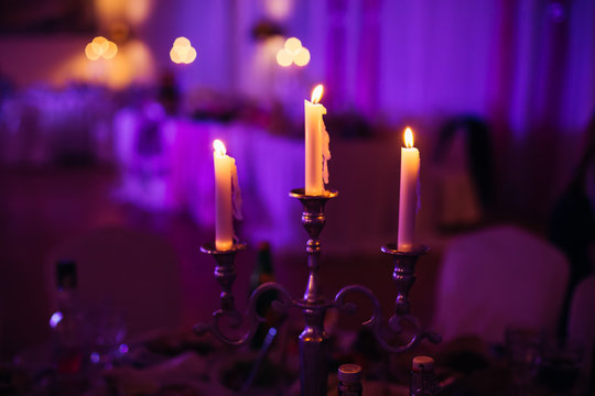 Lighted candles stand on the festive served dinner table in purple light