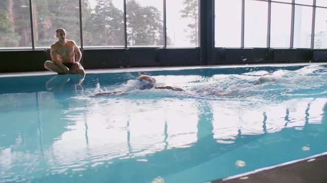 PAN with slow motion of female athlete in goggles and cap swimming with front crawl in indoor pool with blue water while muscular male coach sitting on edge and watching her technique 