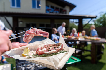 Cropped shot of man preparing meat at outdoor grill and family sitting at table outdoors