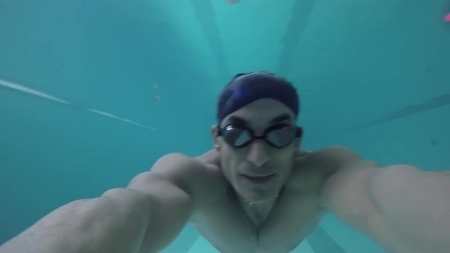 Shot from action camera held by muscular male athlete in goggles and cap swimming underwater in pool, then coming up for air 