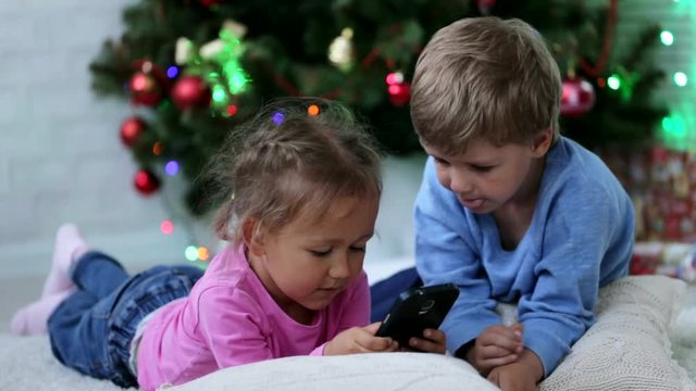 Cute siblings wathing somethng funny in the mobile phone near Christmas tree.