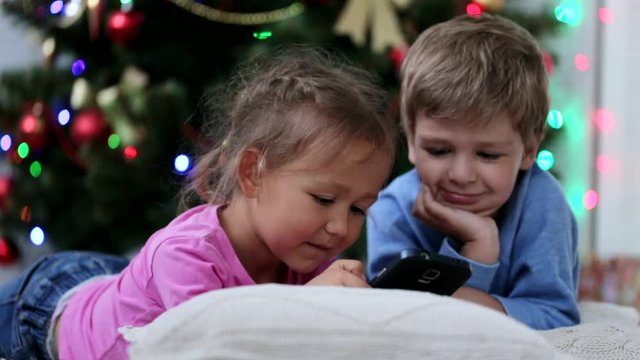 Cute siblings wathing somethng funny in the mobile phone near Christmas tree.