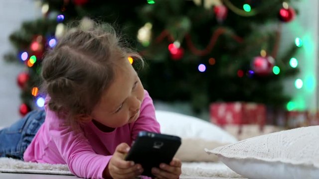Little cute girl using smart phone, in front of Christmas tree. Close-up shot.