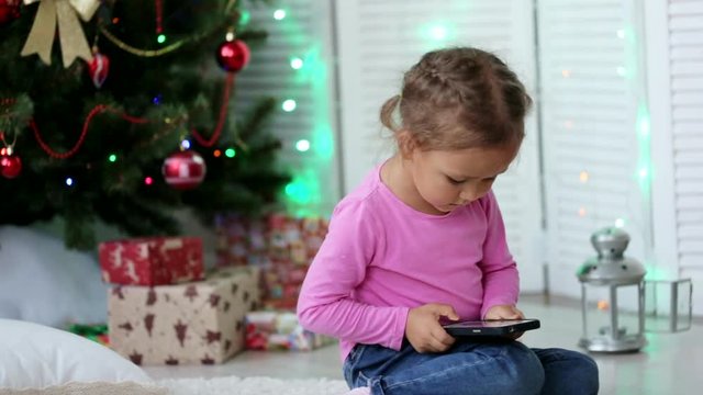 Little cute girl using smart phone, in front of Christmas tree. Close-up shot.