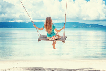 Young woman with long blond hair swinging on a swing on the shore of the tropical sea - obrazy, fototapety, plakaty