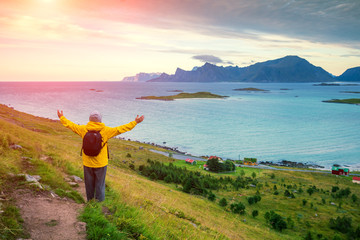 Panoramic aerial view of the sea at sunset. Man tourist with hands in the air standing on a cliff of rock. Beautiful mountain landscape. Nature Norway, Lofoten Islands