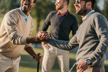 happy multicultural friends shaking hands while playing golf on golf course