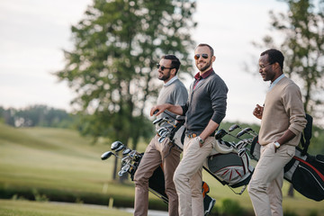 stylish multiethnic men holding bags with golf clubs and walking on golf course