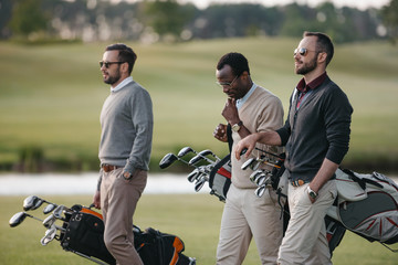 Multiethnic golfers holding bags with golf clubs and walking on golf course