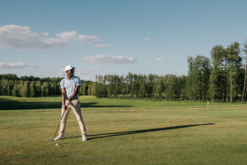 Full length view of man in cap holding golf club and hitting ball on green lawn