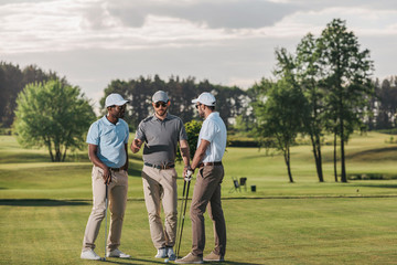Multiethnic group of golfers holding clubs and talking while standing on green grass