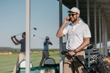 smiling golf player talking on smartphone while friends playing golf