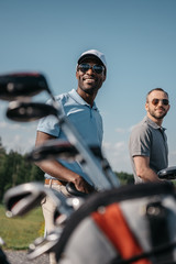 smiling multiethnic sportsmen going to the golf course, bag with clubs on foreground