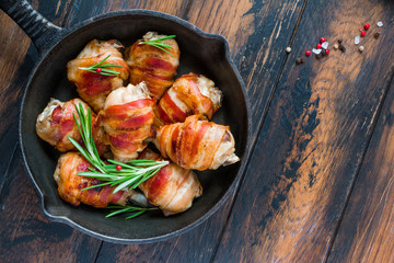 Bacon wrapped chicken drumsticks in a black cast-iron skillet on the wooden rustic table, top view.