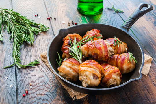 Bacon wrapped chicken drumsticks in a black cast-iron skillet on the wooden rustic table.