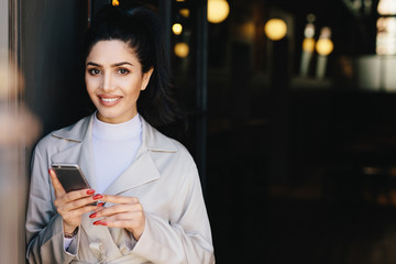 Attractive brunette businesswoman having dark hair tied in pony tail and wonderful make-up smiling demonstrating her white perfect teeth waitng for her business partner using modern smartphone