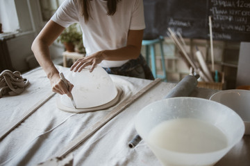 Sculptor cuts grey raw clay round plastic bowl for modeling ceramic plate on big table with industrial fabric in workshop