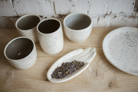 Stylish chamotte ceramic hand with dry lavender and handmade cups on wooden shelf with white brick wall on background in workshop