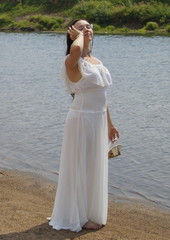 Fototapeta na wymiar Brunette girl in white light dress barefoot on the shore of a small river