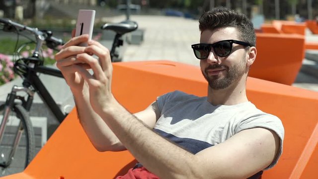 Handsome man sitting on the orange seat in the city and doing selfies on smartphone, steadycam shot
