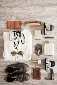 Preparation for traveling concept, dress, old shoes, jeans, beige hat, vintage cameras, leather bag, sunglasses, wallet, woman bra and another stuff on a white wooden background.