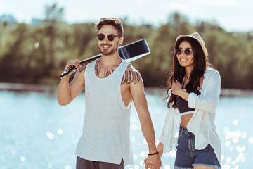 smiling interracial couple in sunglasses holding hands while man carrying guitar