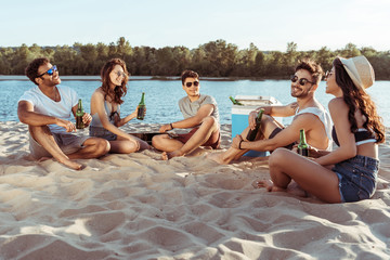 happy young friends drinking beer while relaxing on sandy beach at riverside