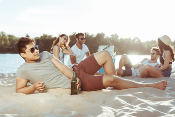 young man lying on sandy beach while her friends resting behind on riverside