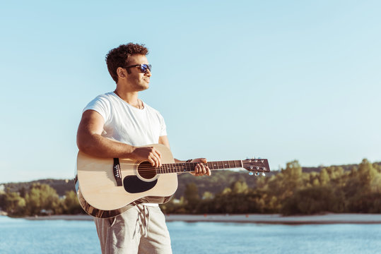 young african american man playing guitar and looking away while standing on riverside