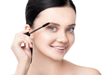 naked brunette woman applying mascara and smiling at camera isolated on white