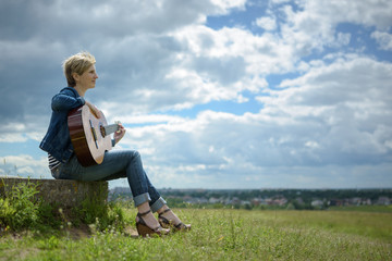 Obraz premium young woman with short blond hair sitting with guitar on the field in cloudy day