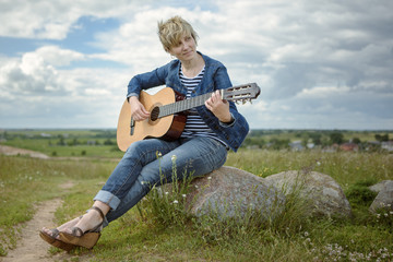 Young woman playing guitar sitting on rock in the field in windy weather