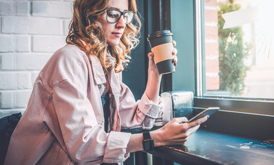 Hipster girl sitting in cafe at black table, drinking coffee and using smartphone. Businesswoman...