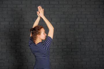 The girl is practicing meditation in yoga class against a gray brick wall. Young slim sporty girl holds her hands over head in Namaste posture! Woman do yoga indoors.