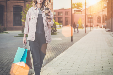 Young woman in glasses is walking along city street,carrying shopping bags and holding cup of coffee.