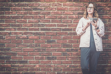 Young attractive woman in glasses, pink denim jacket stands outside on red brick wall background and uses smartphone.