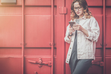 Young woman in glasses stands outside on red metal wall background,looking at screen of phone,uses...