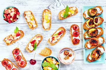 Top view of assorted crostini (toasts) with cut fresh fruits: cherry, peach, pear and grilled figs on white wooden table.