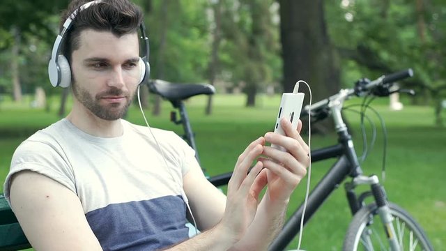 Handsome man listening music in the park and doing selfies on smartphone, steadycam shot
