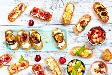 Small toasts (crostini) with cream cheese and cut fresh fruits (pear, peach, cherry, grilled figs) on white table. Top view of summer party appetizers.