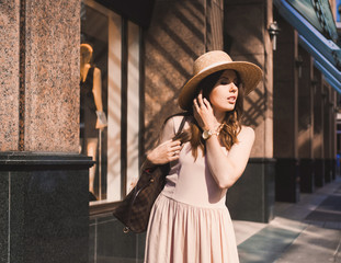 Close-up portrait of a slender young girl blogger beautiful brunette in downtown dusseldorf in a pastel dress and a lady's hat wearing sunglasses walking poses and smiling on the sunset