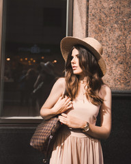 Close-up portrait of a slender young girl blogger beautiful brunette in downtown dusseldorf in a pastel dress and a lady's hat wearing sunglasses walking poses and smiling on the sunset