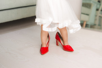 Cute funny little baby girl walks at home trying mom's red high heel shoes